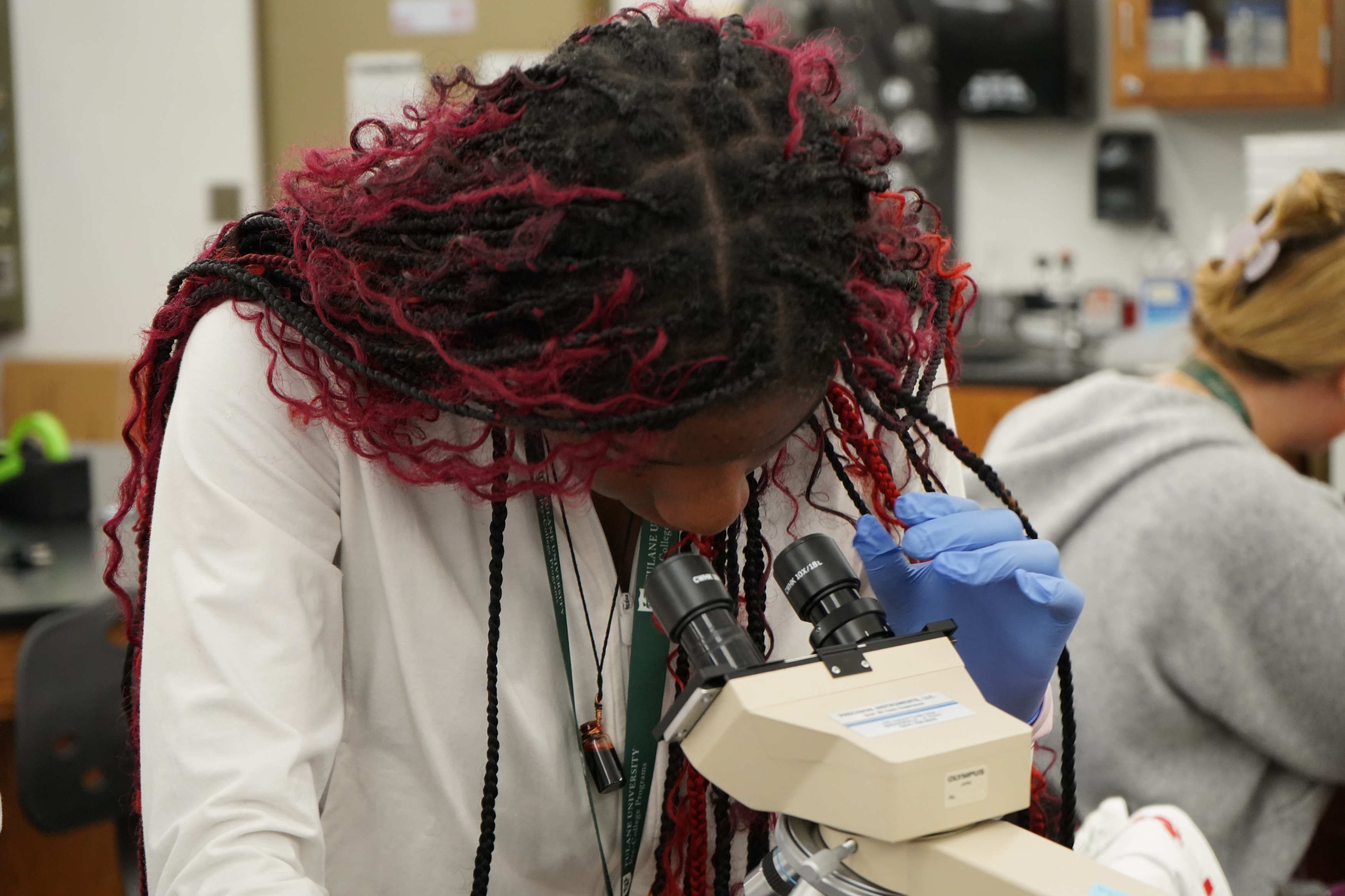Student with red-streaked braids peers into a microscope in a science lab.