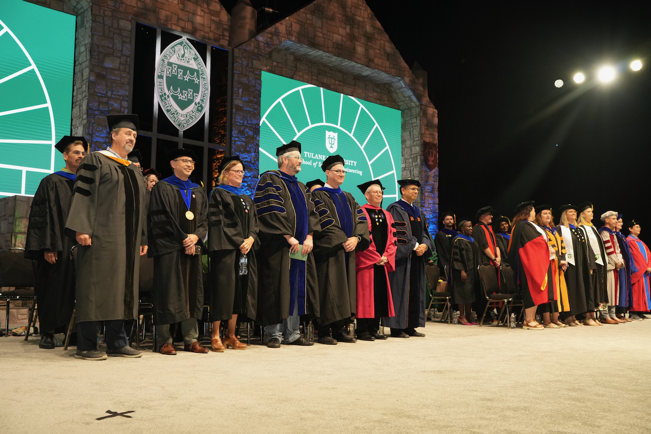 Photograph of graduation faculty in academic regalia standing on a stage.