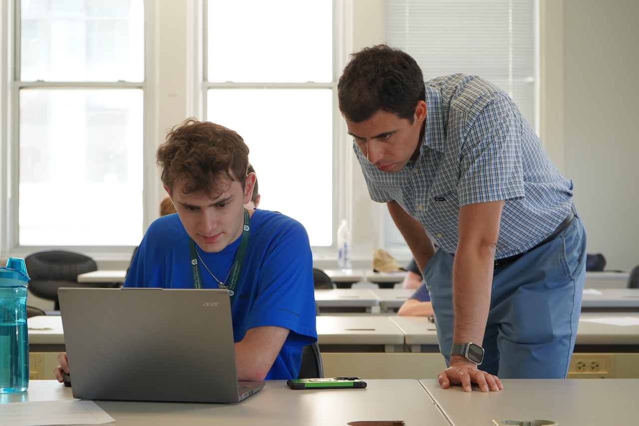 A photograph of a teacher helping a student on a laptop in a classroom.