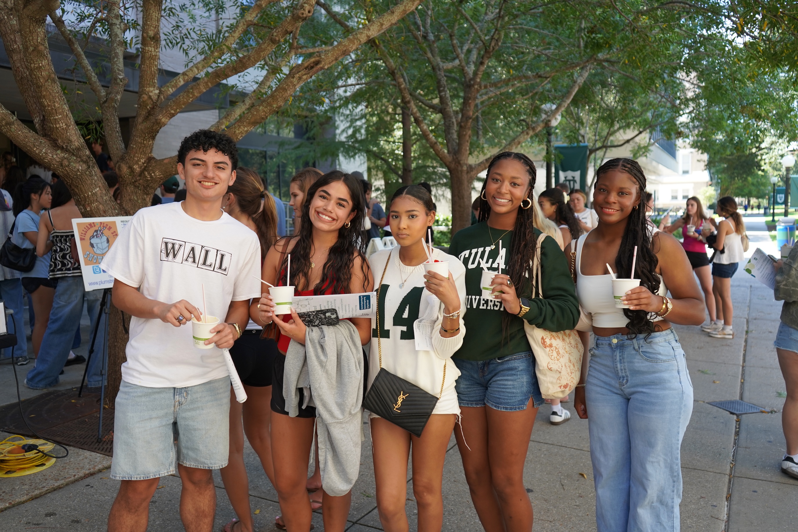 Four young adults stand together smiling, holding drinks outdoors.