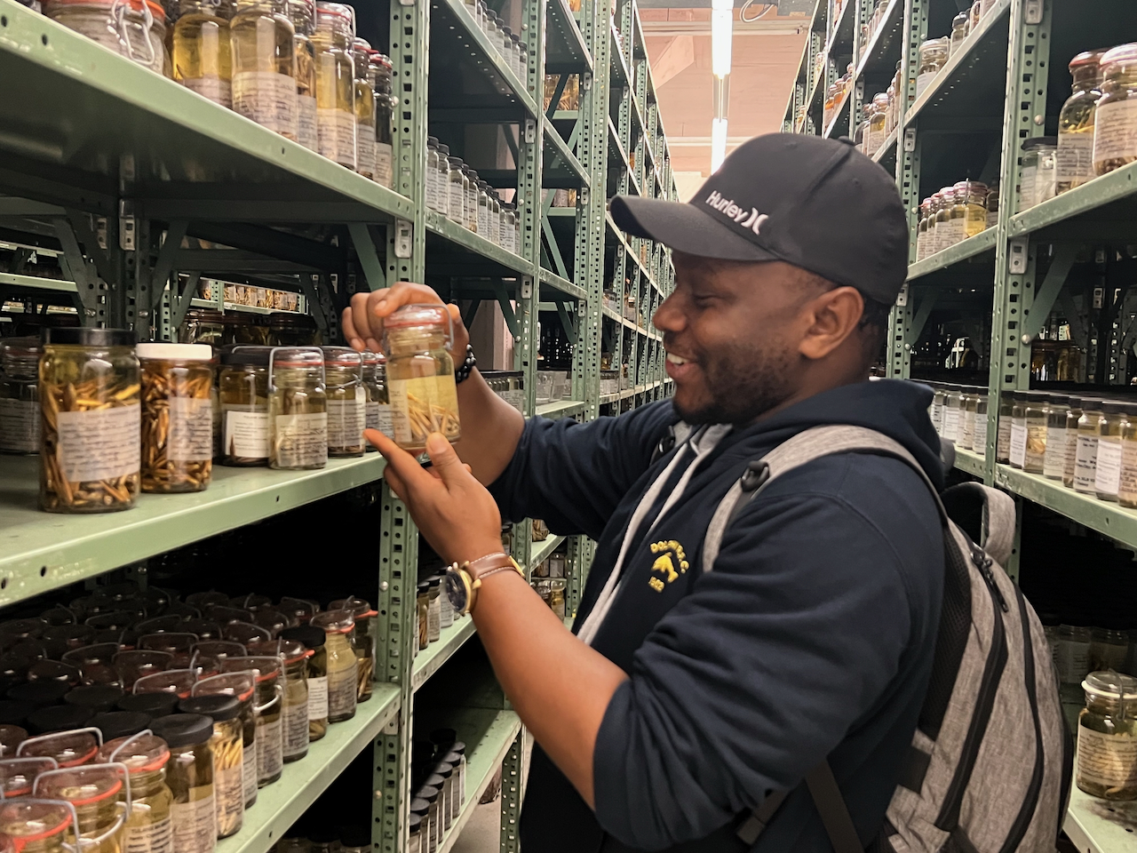 A Black man wearing a cap and backpack examines a jar in a warehouse with tall shelves.