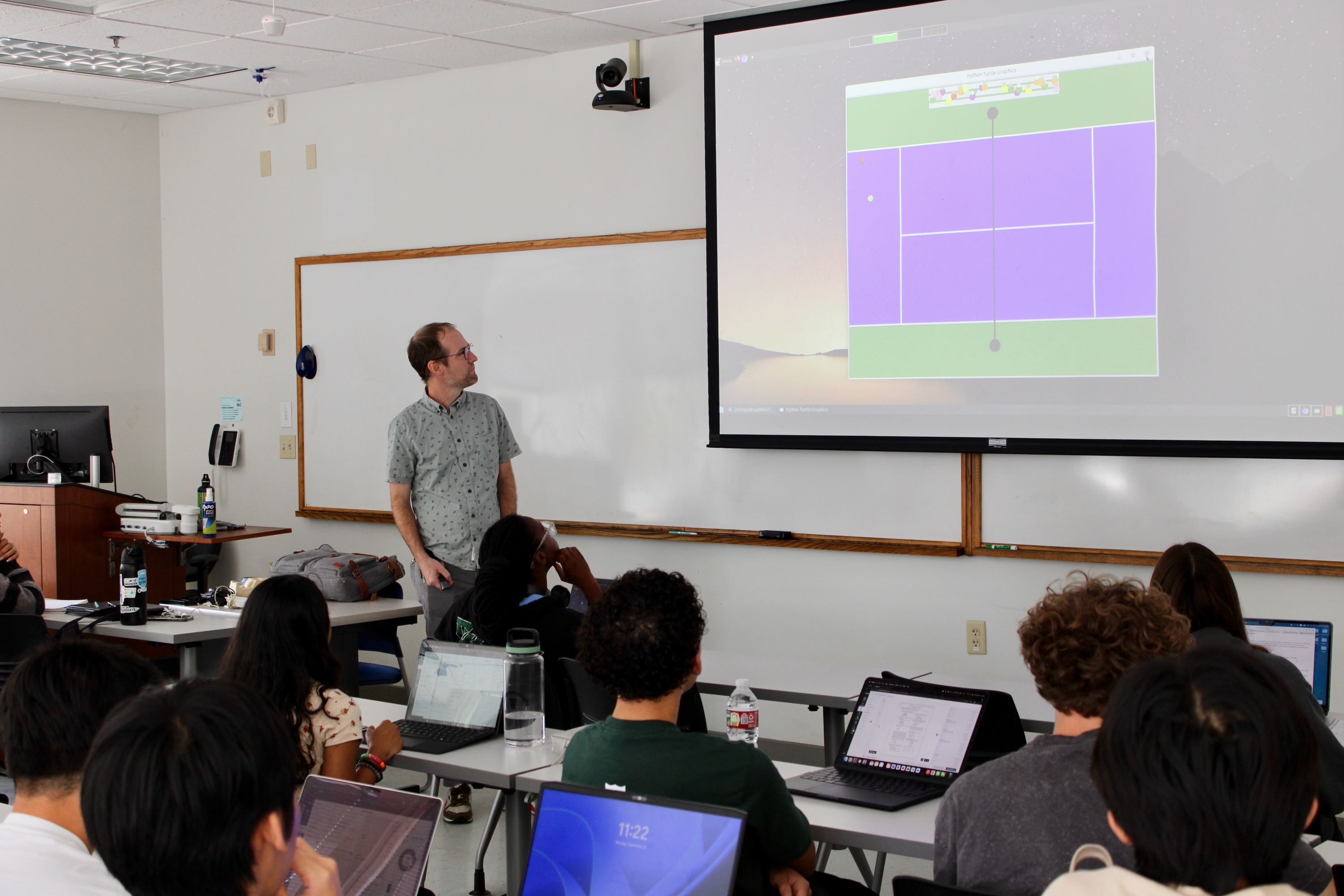 Man presents a screen showing a game interface to students in a classroom.