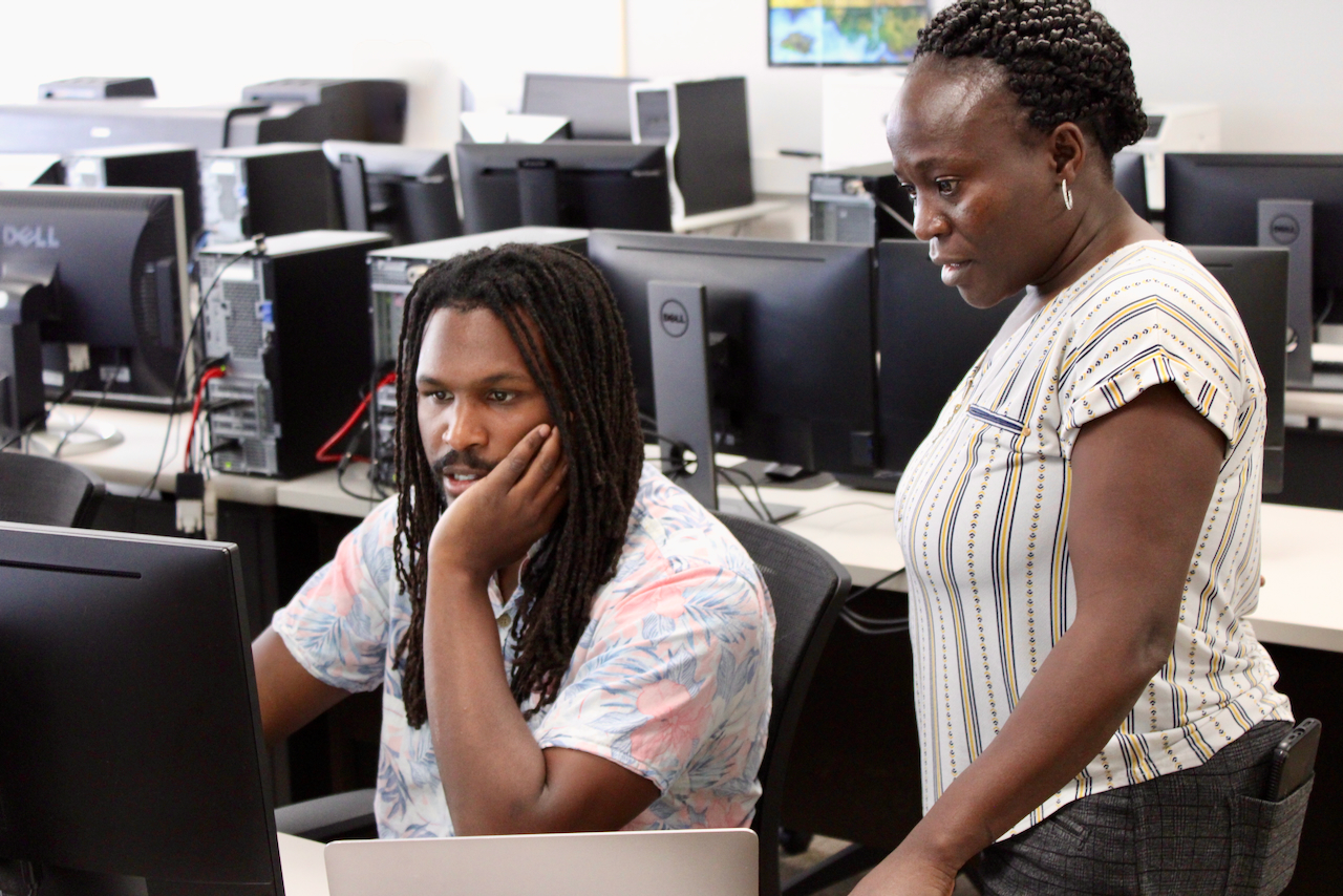 A Black man with dreadlocks looks at a computer screen while a Black woman with her hair in a bun stands beside him.