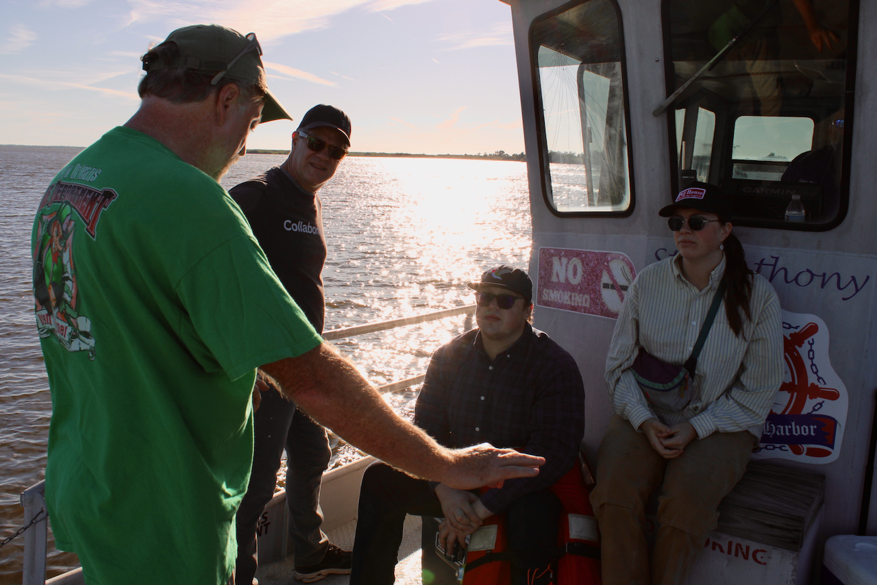 Four people on a boat deck, with a man in green speaking to three others.
