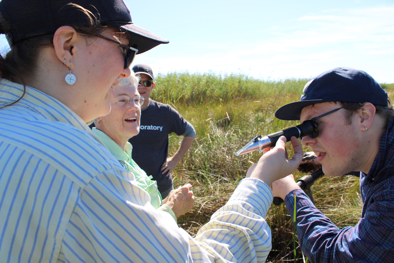 Young man uses a handheld device to examine water with three people watching in a grassy field.