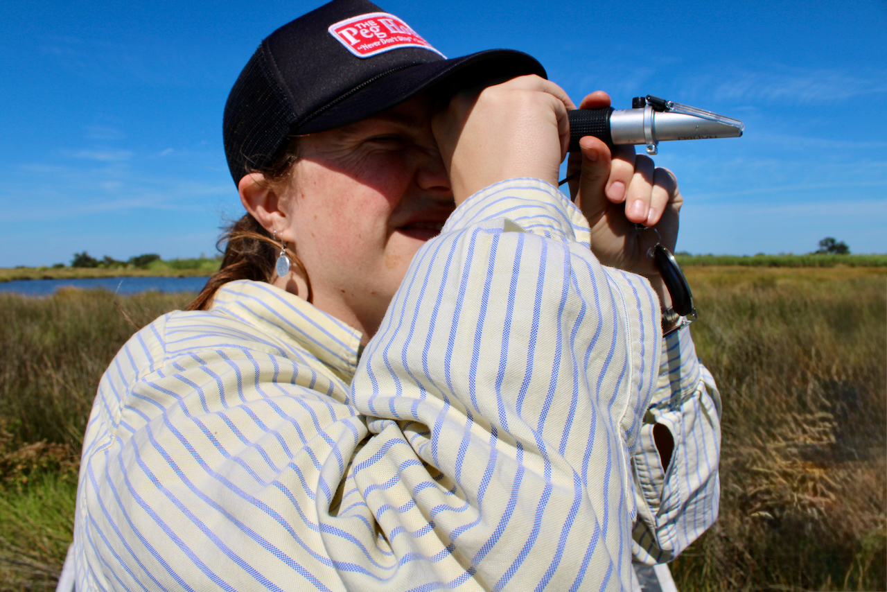 Photograph of a woman in a wetland looking through a refractometer against a blue sky.