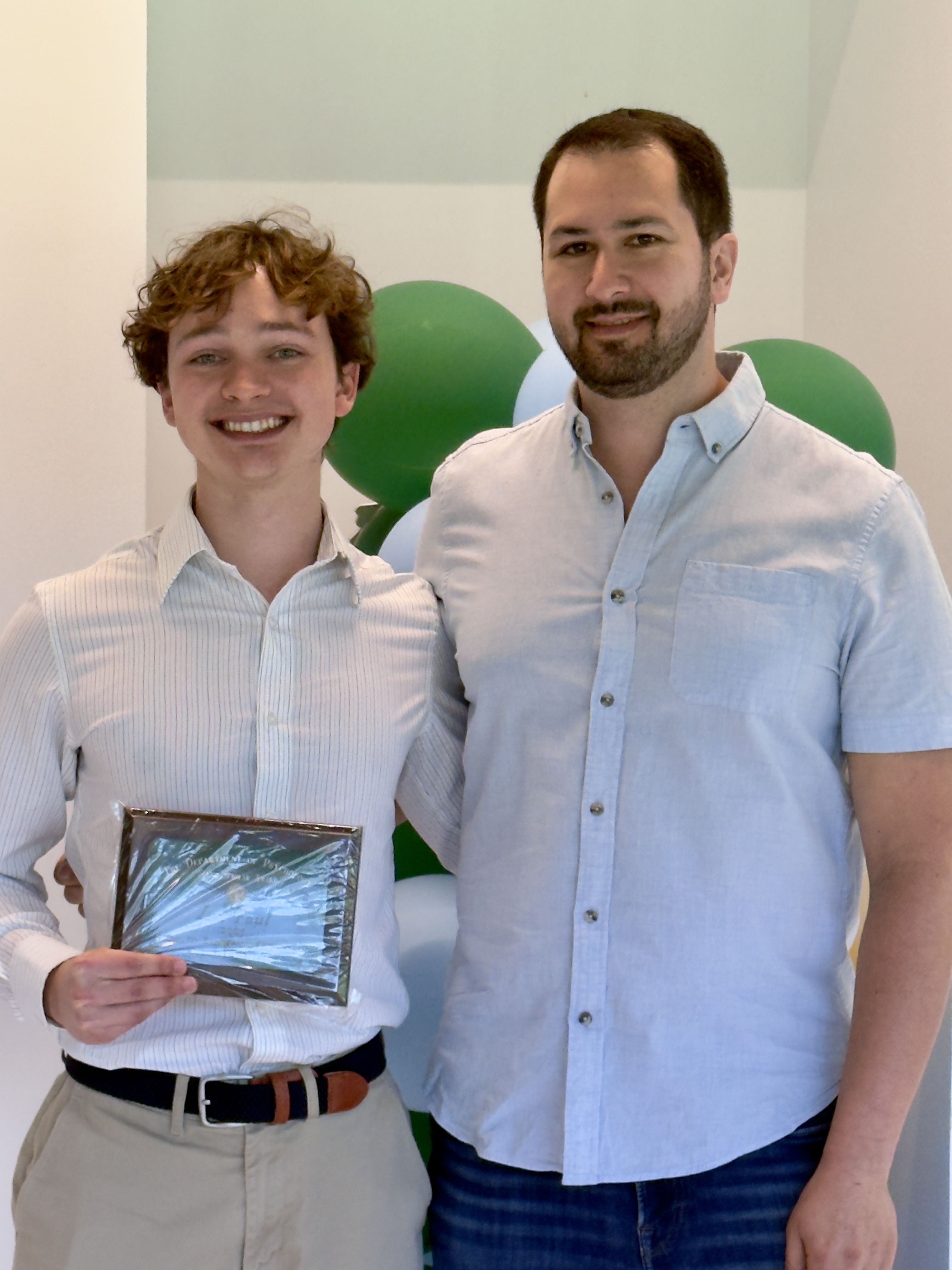 Photograph of two smiling men in light shirts holding a plaque indoors.
