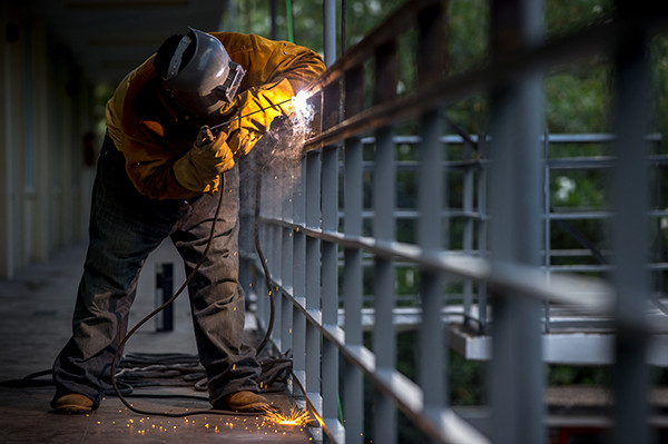 Welder in a yellow jacket sparks metal railing outdoors.