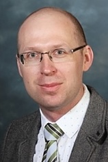 A man wearing glasses and a suit smiles at the camera.