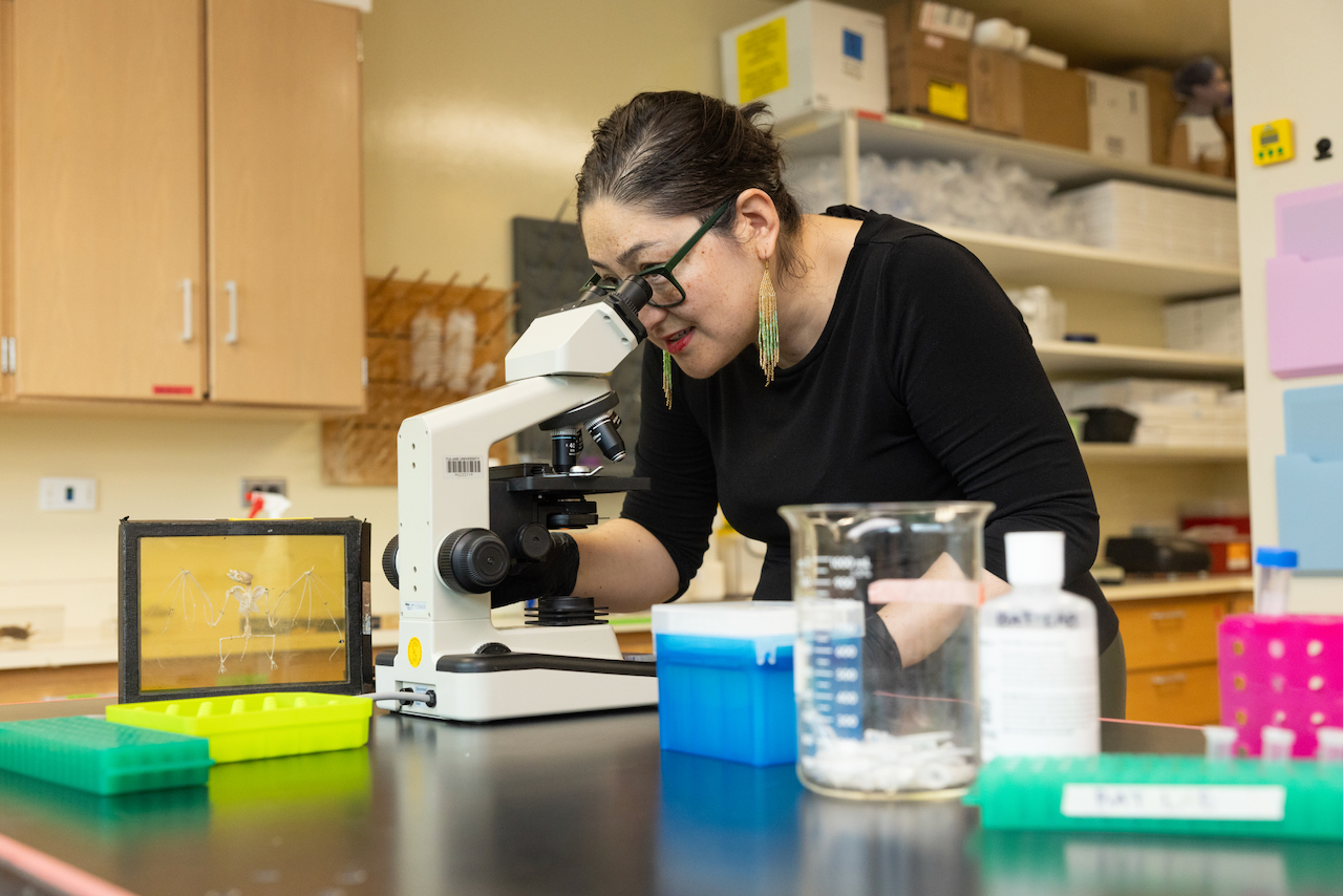 Woman with glasses and black gloves peers into a microscope in a lab.