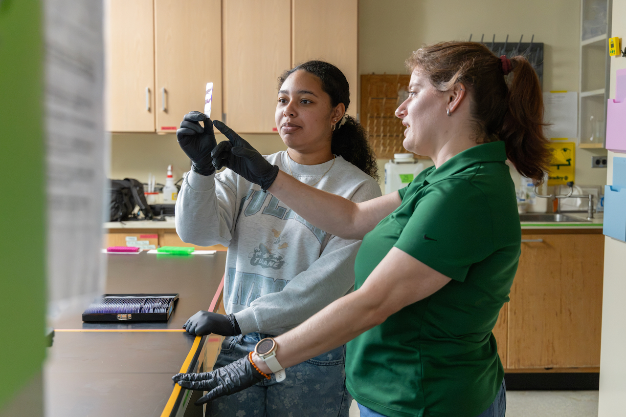 Two women in a lab, one pointing to a slide in gloved hands.