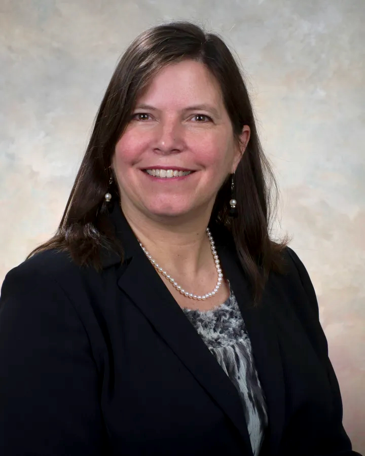 Photograph of a smiling woman with dark hair wearing a black jacket and a pearl necklace.