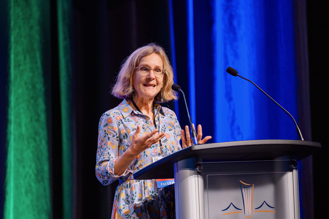 Photograph of a smiling woman speaking behind a podium on a stage with blue curtains.