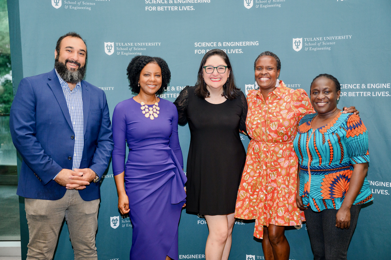 Five individuals pose for a photograph, smiling against a backdrop with university logos.