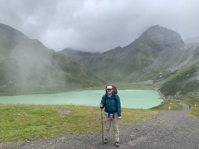Hiker with backpack and trekking pole stands before a turquoise lake and misty mountains.