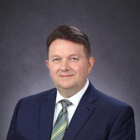 Photograph of a smiling middle-aged man in a dark suit and striped tie against a gray background.