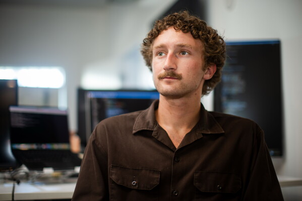 Photograph of a young man with curly brown hair and a mustache in an office.