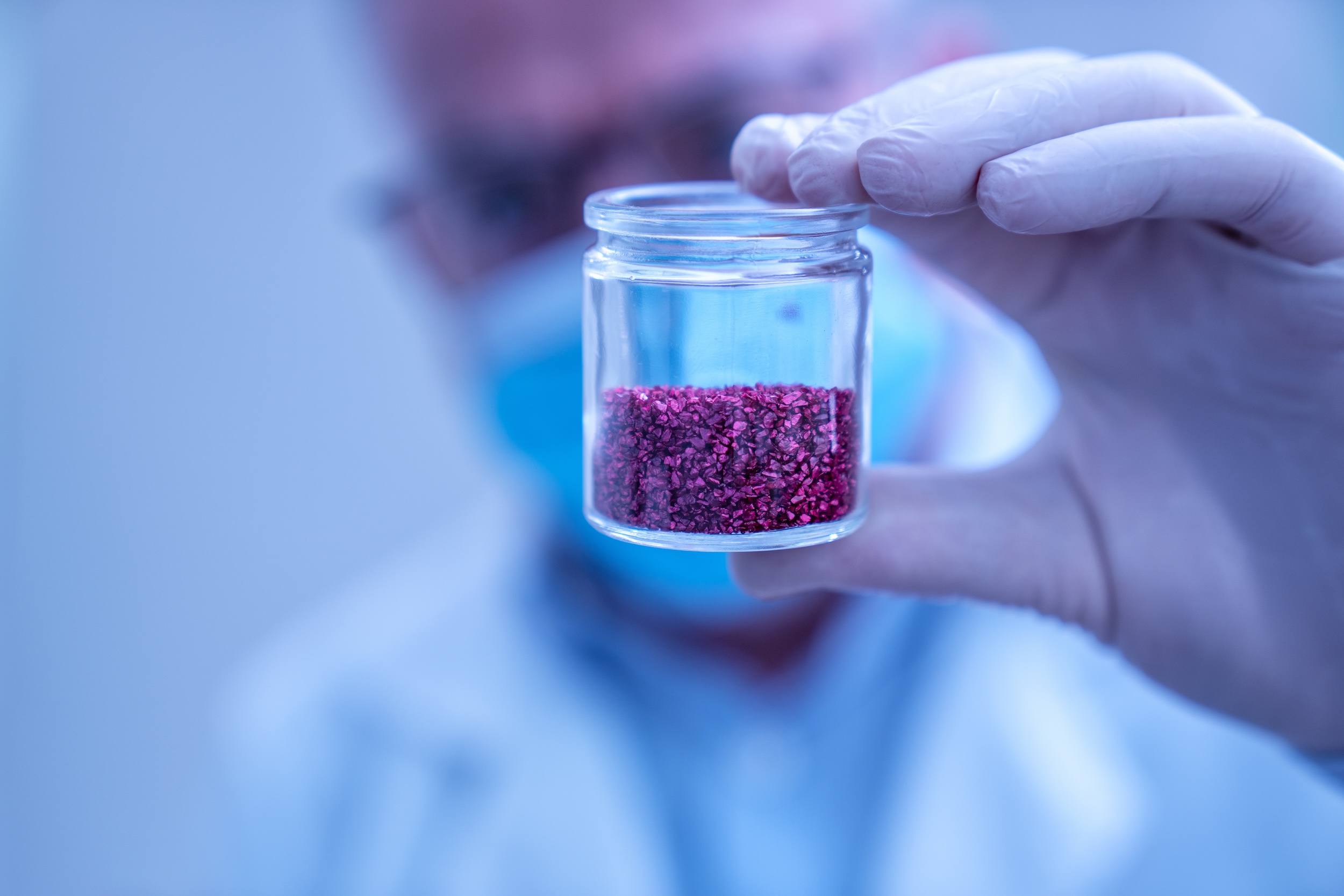 Man in lab coat holding a clear glass jar with grains of red material in it