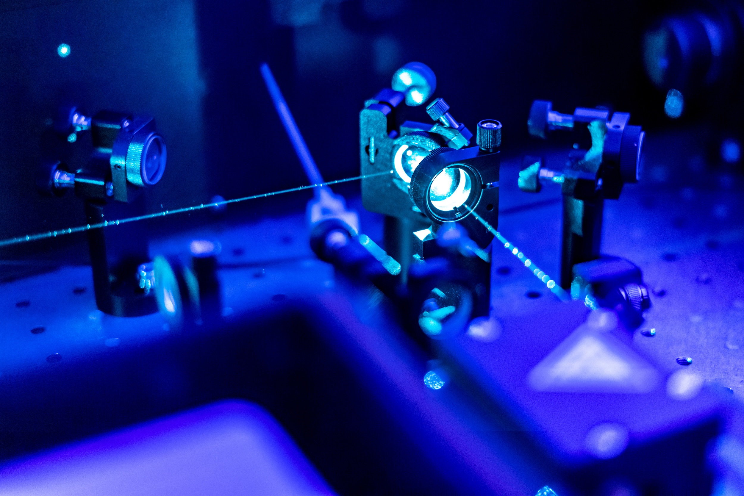 Photograph of an optical physics lab bench with blue laser beams passing through lenses and mirrors.