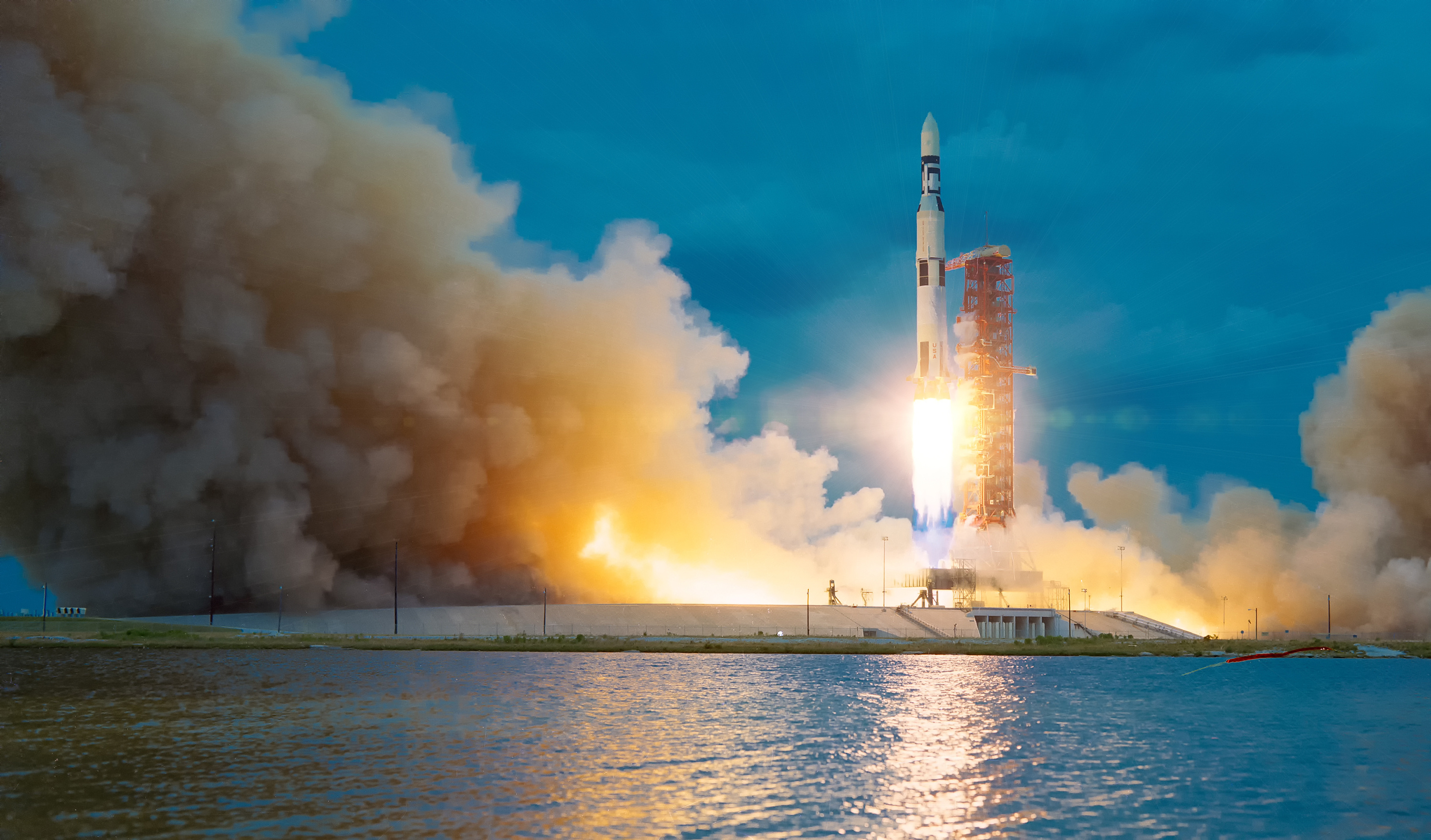 Photograph of a rocket launching near water, producing a massive cloud of smoke and fire.