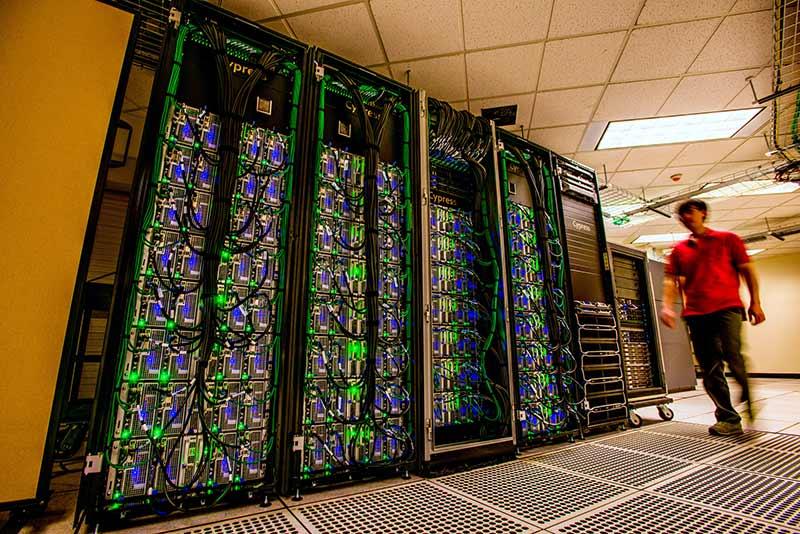 Photograph of a person walking past server racks glowing with green and blue lights.
