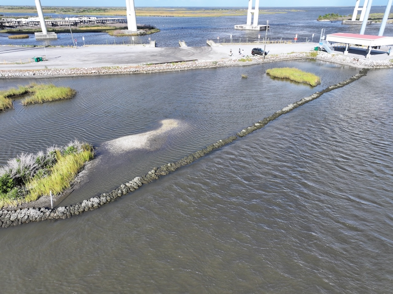 Aerial photograph of a marshland with a rock barrier in the water and bridge pillars in the background.
