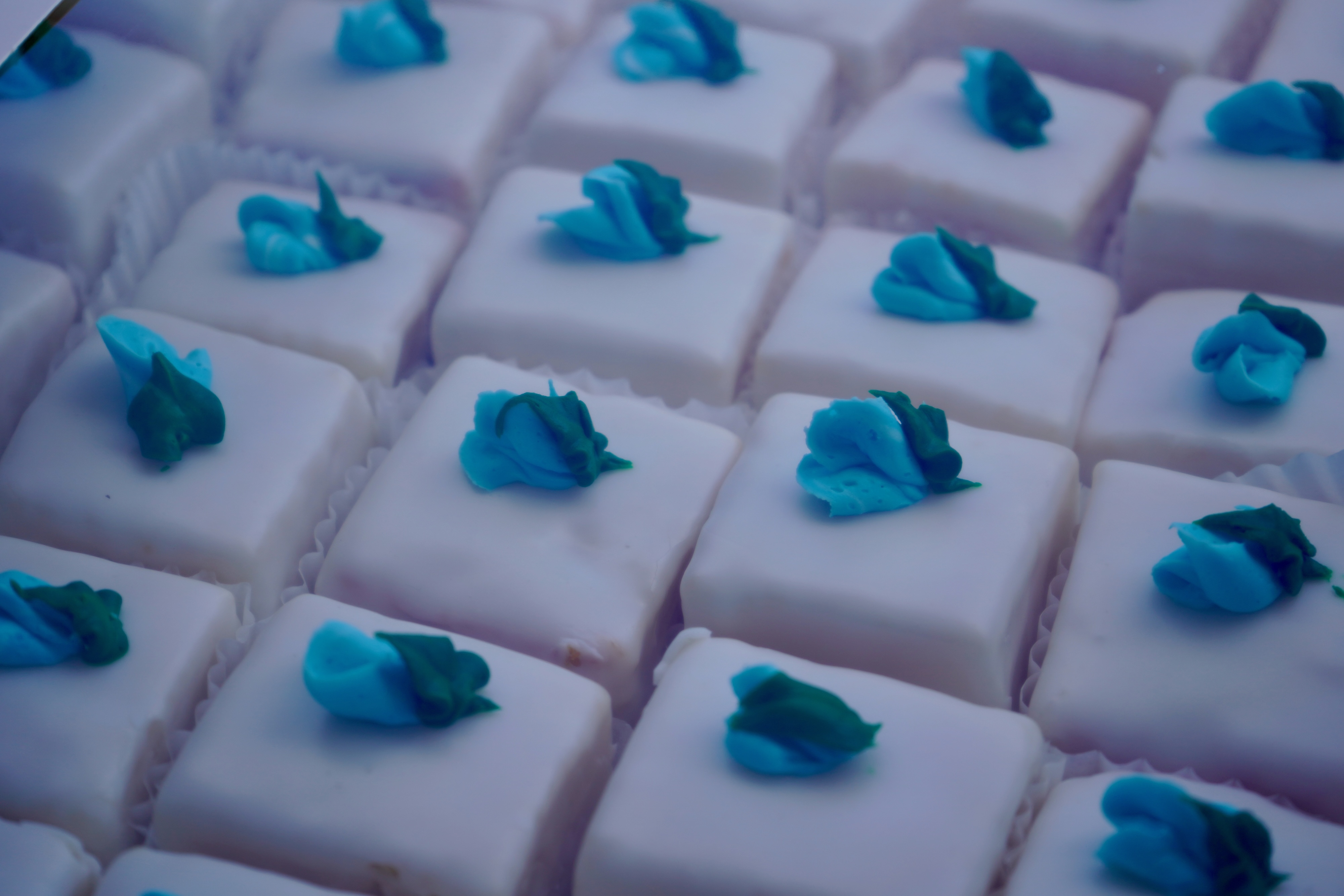 Close-up photograph of rows of small white cakes with blue and green frosting decorations.