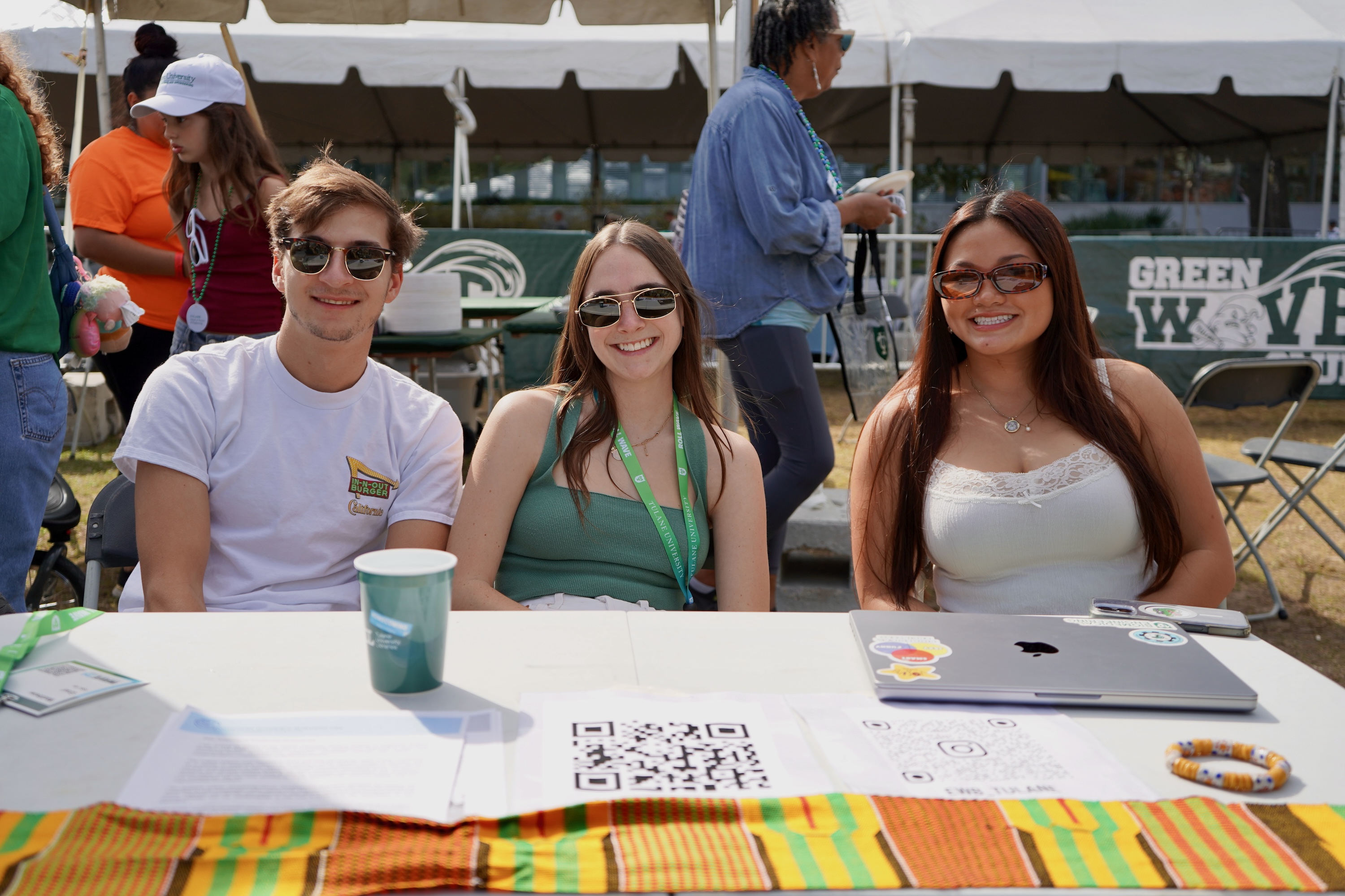 Photograph of three smiling students sitting behind a table outdoors.