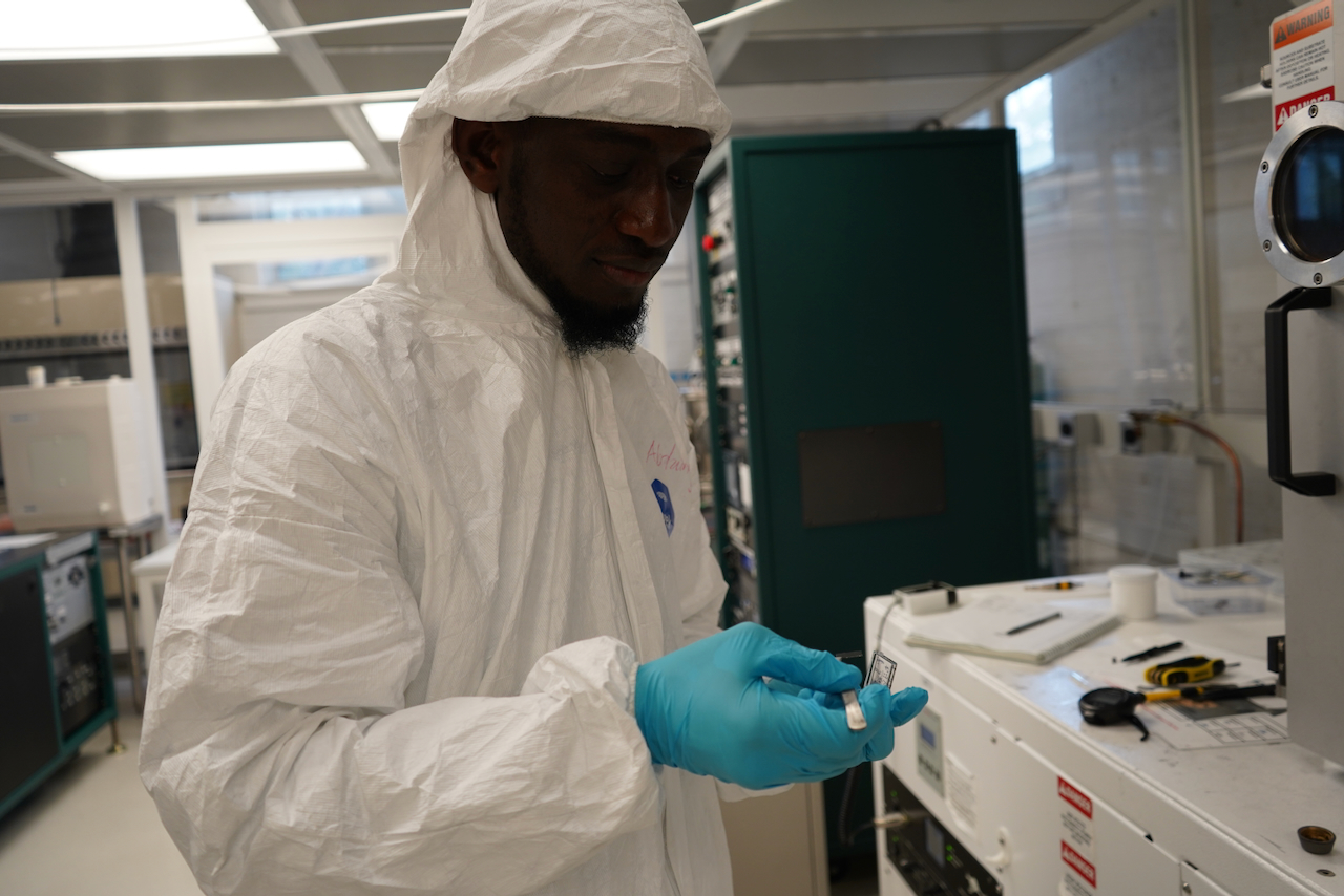 Photograph of a scientist wearing a cleanroom suit and blue gloves in a lab.