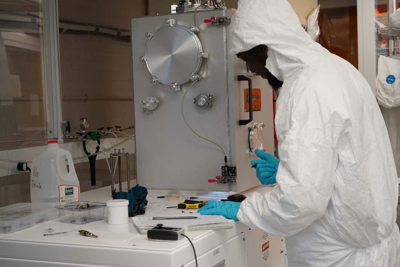 A researcher in a white hazmat suit and blue gloves works at a lab bench.