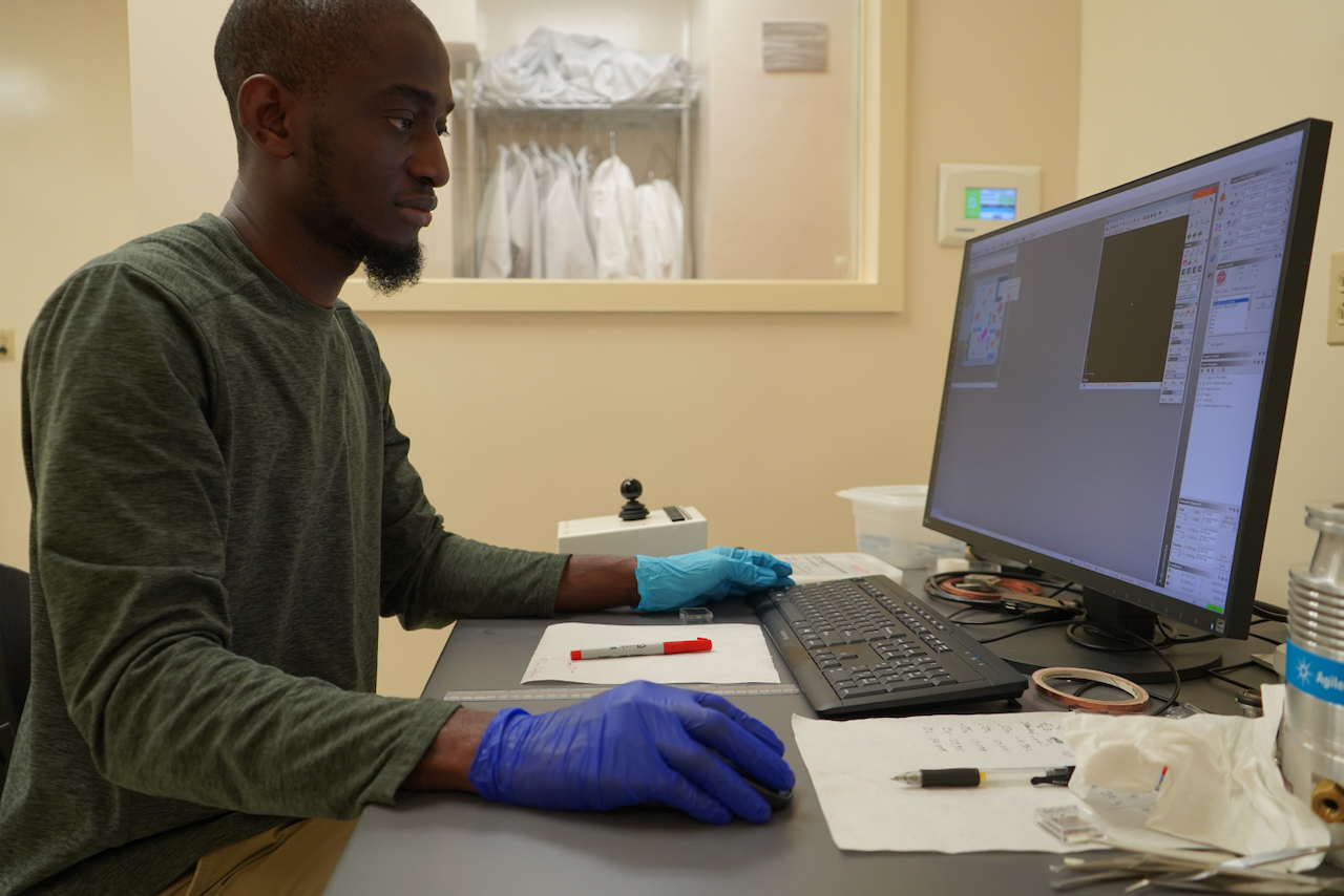 A Black man wearing blue gloves works on a computer in a brightly lit room.