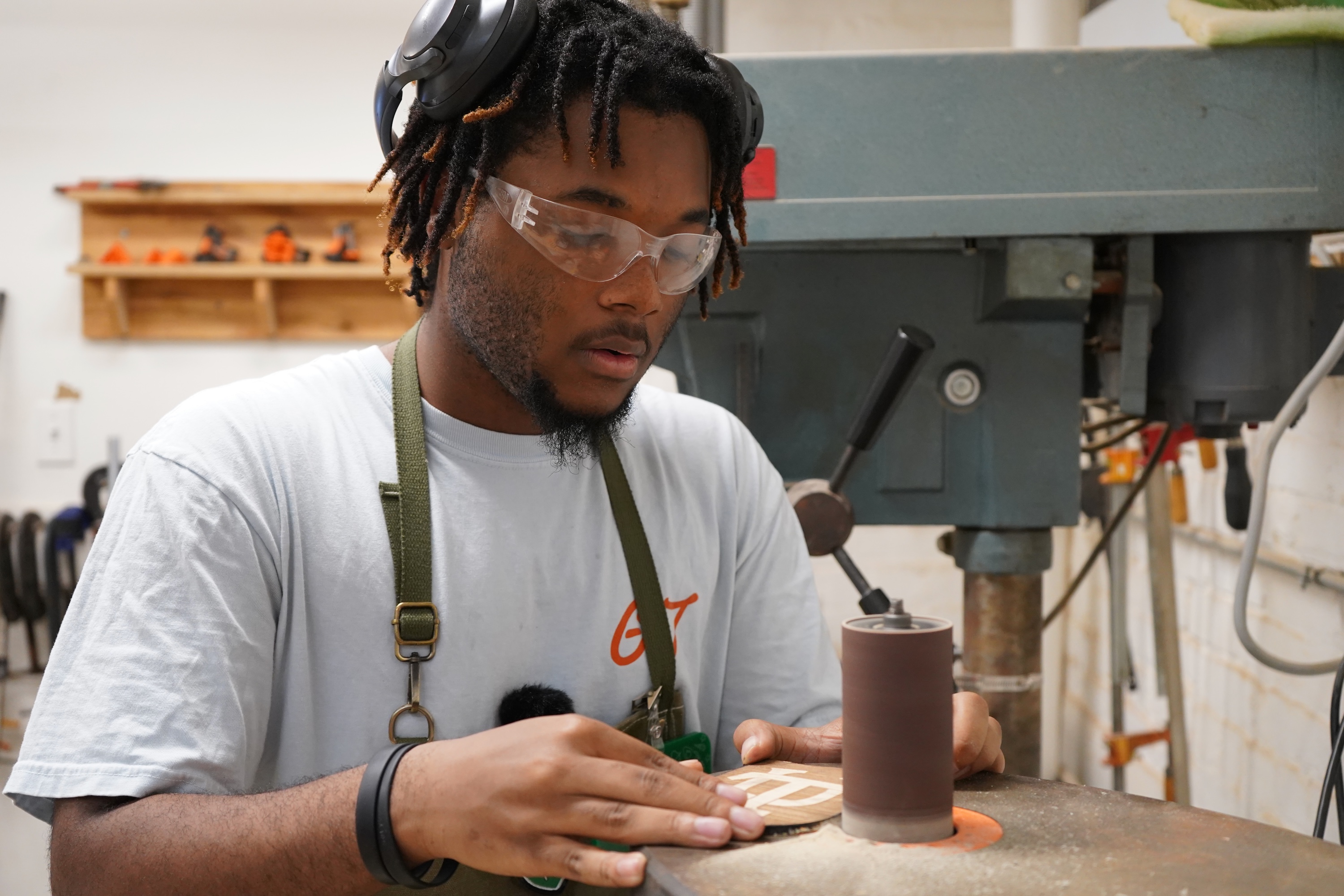 Photograph of a Black man wearing safety glasses using a drill press in a workshop.