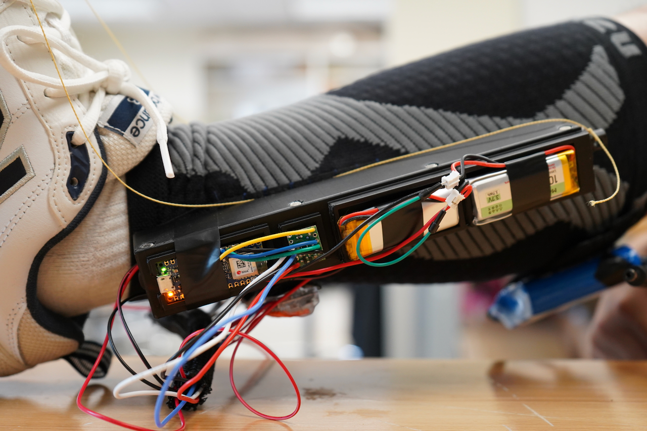 Photograph of a wearable electronic device strapped to a leg, with exposed wires and batteries.