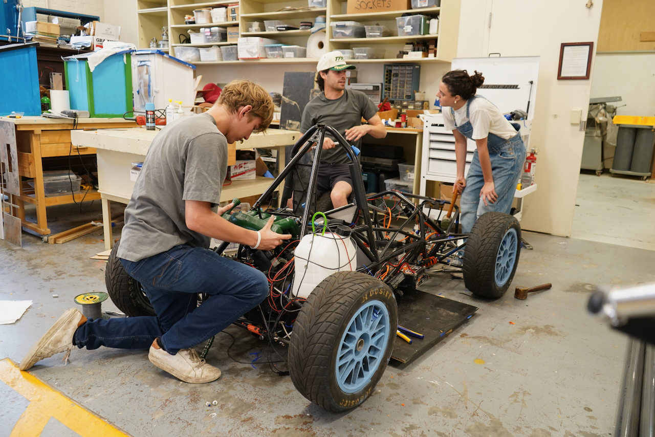 Photograph of three students working on a race car frame in a workshop.