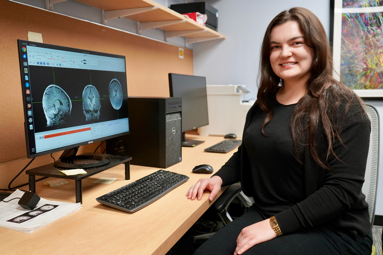 A smiling woman seated at a desk with dual monitors displaying brain scans.