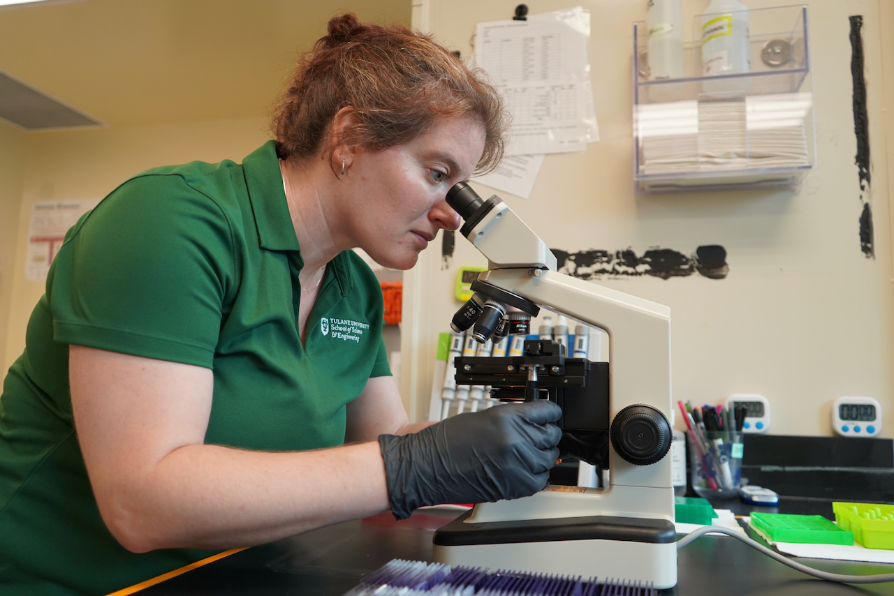 Woman wear a green polo and black gloves looking into a microscope