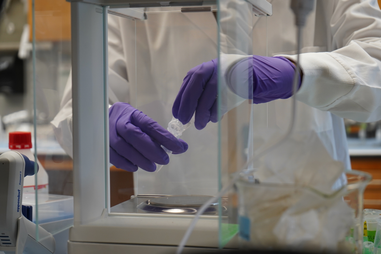 A scientist wearing purple gloves holds a small vial inside a lab enclosure.