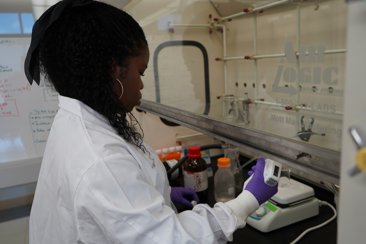 Photograph of a Black female scientist in a lab coat working inside a fume hood.