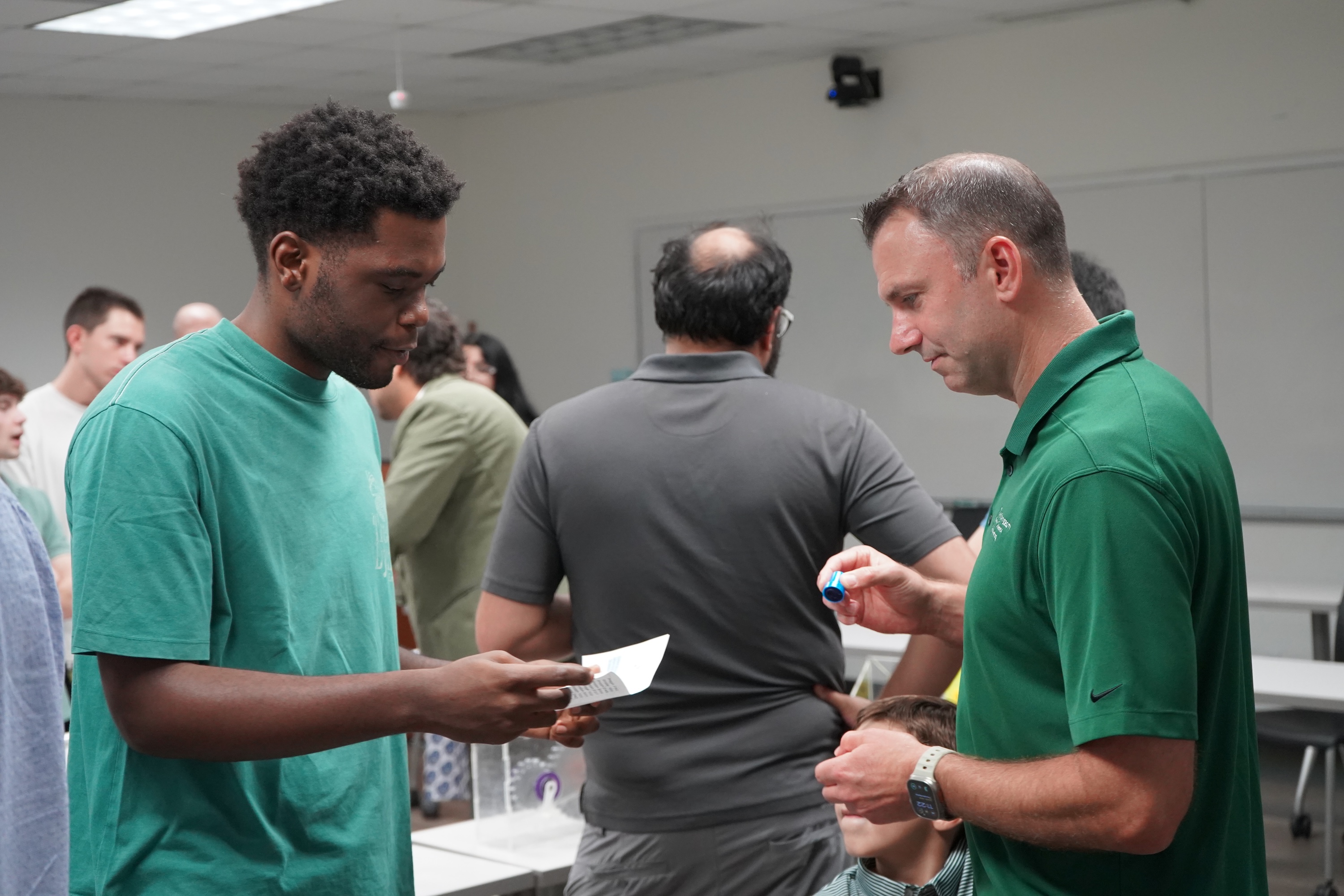 Photograph of a Black man and a Caucasian man talking while holding papers in a room with other people.