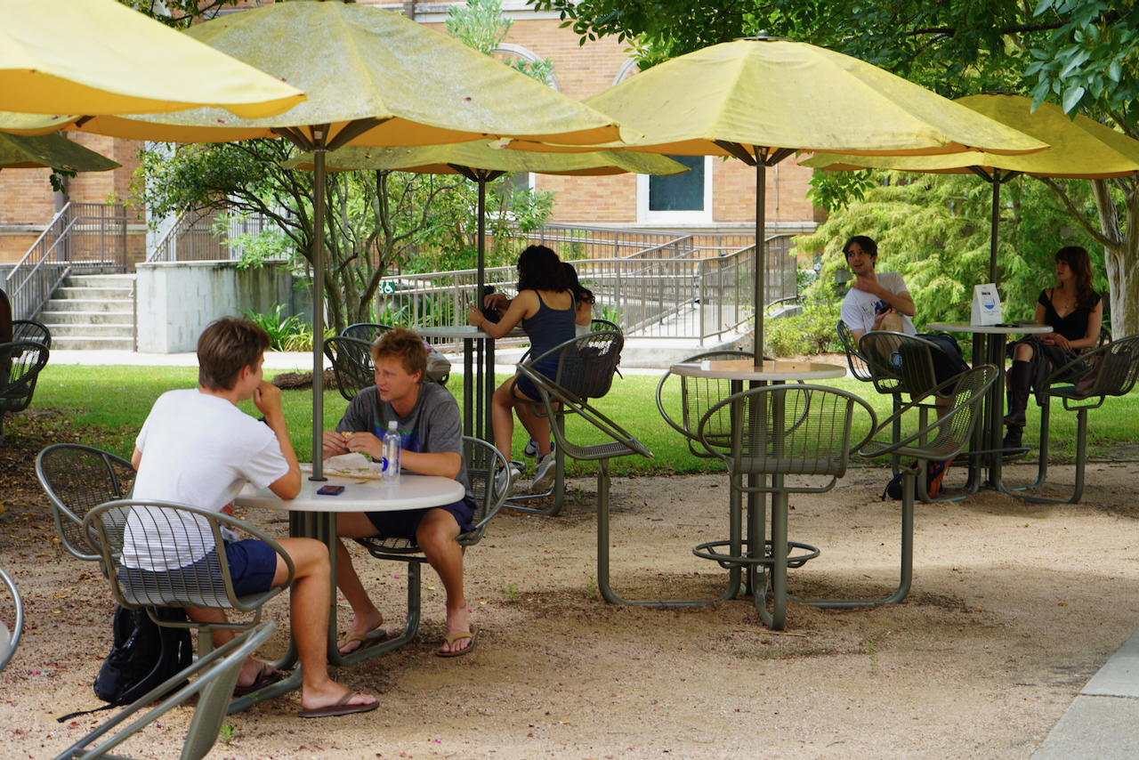 Photograph of students sitting at outdoor tables under yellow umbrellas on a sunny day.