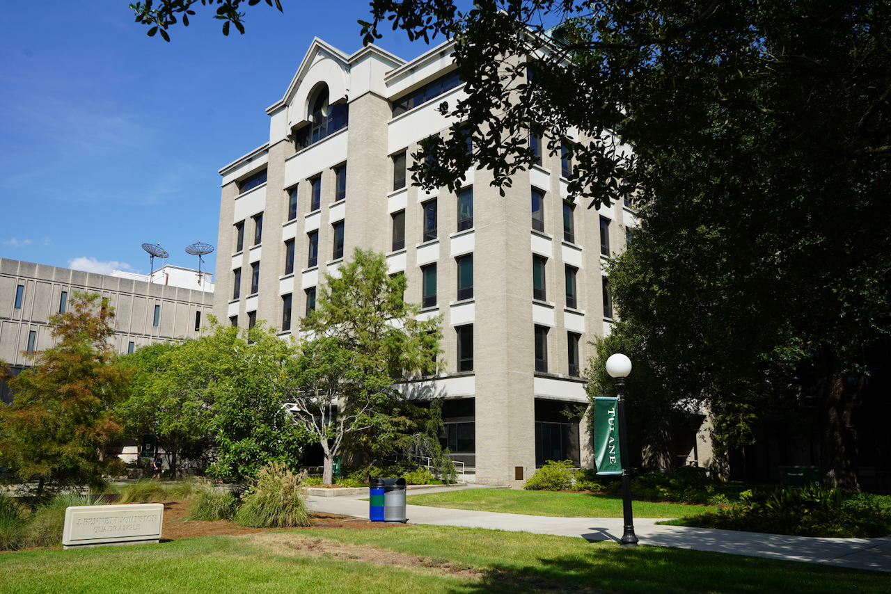 Photograph of a multi-story campus building surrounded by trees under a blue sky.