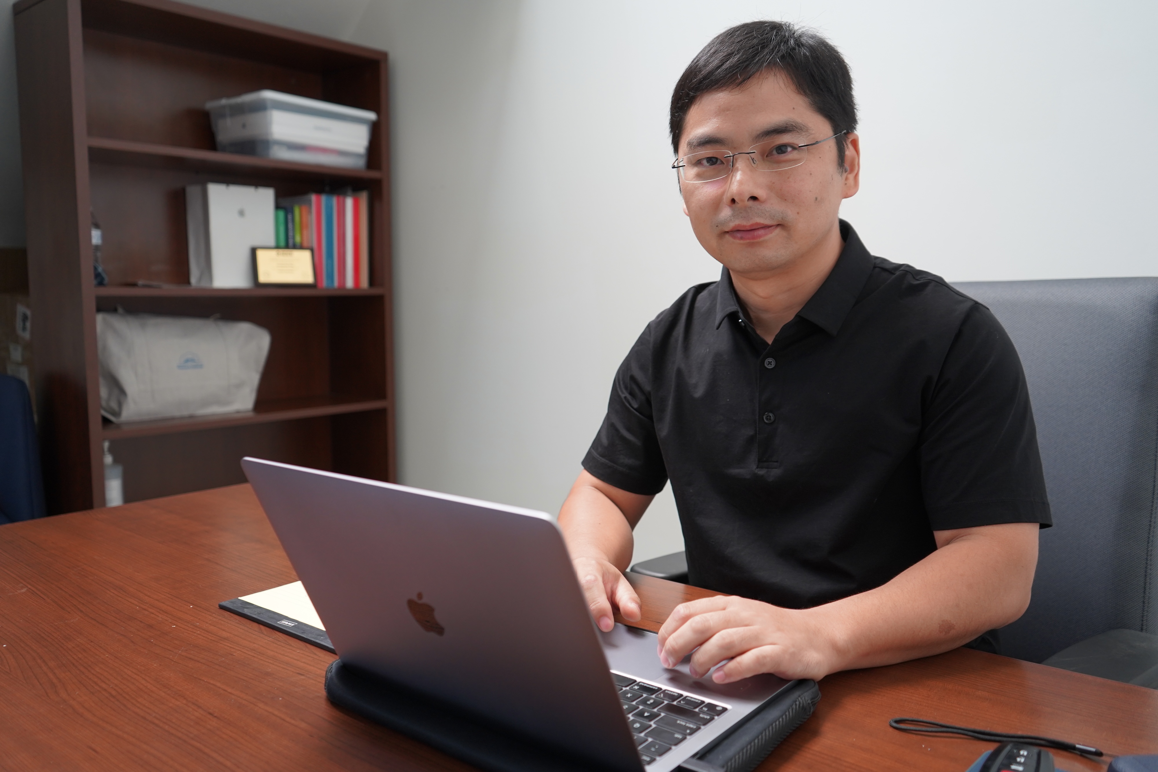 A photo of a man sitting at a desk with a laptop, looking at the camera.