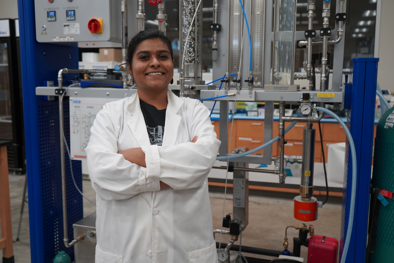 Photograph of a smiling female researcher in a lab coat standing before complex laboratory equipment.