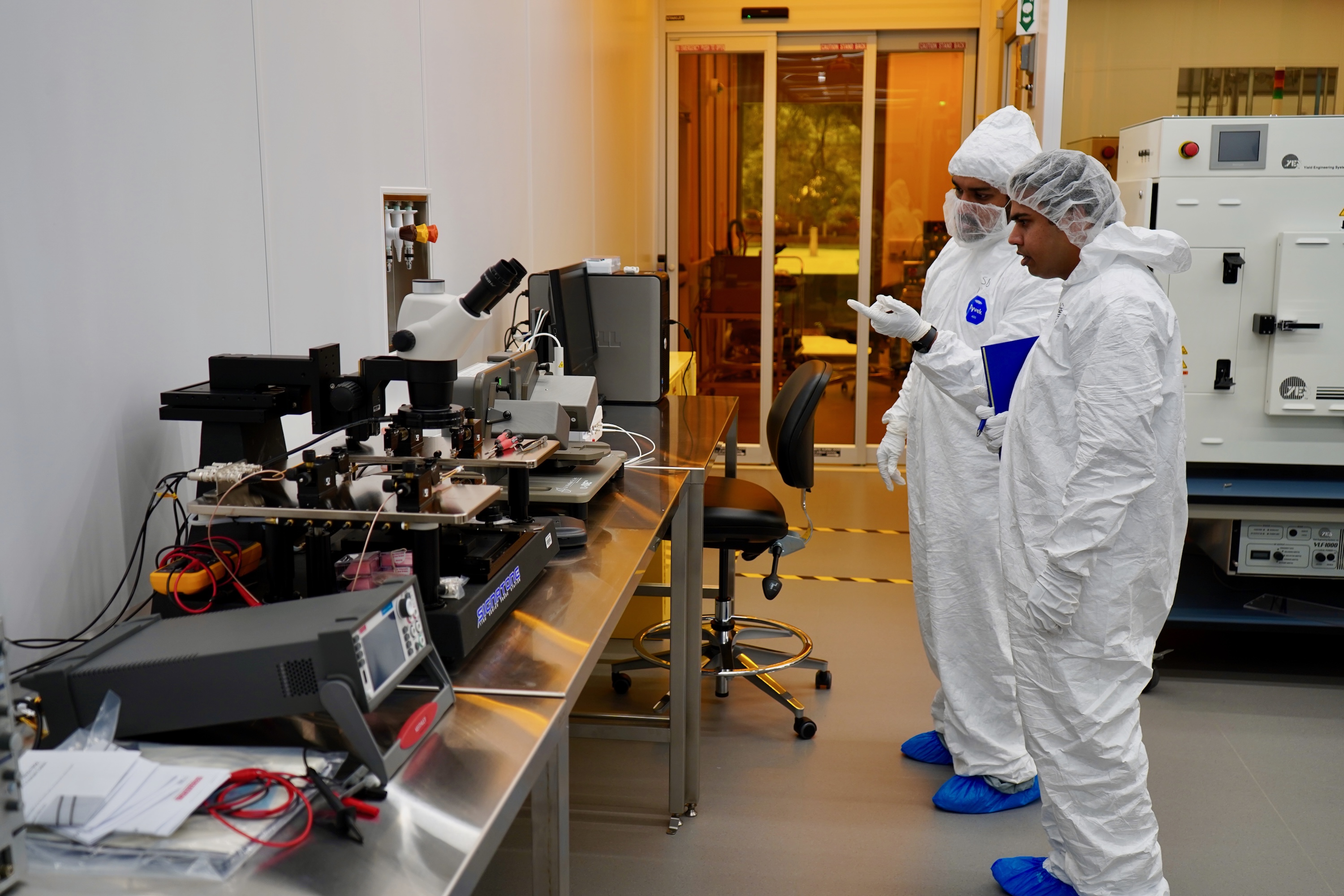 Two technicians in cleanroom suits review equipment in a brightly lit laboratory.
