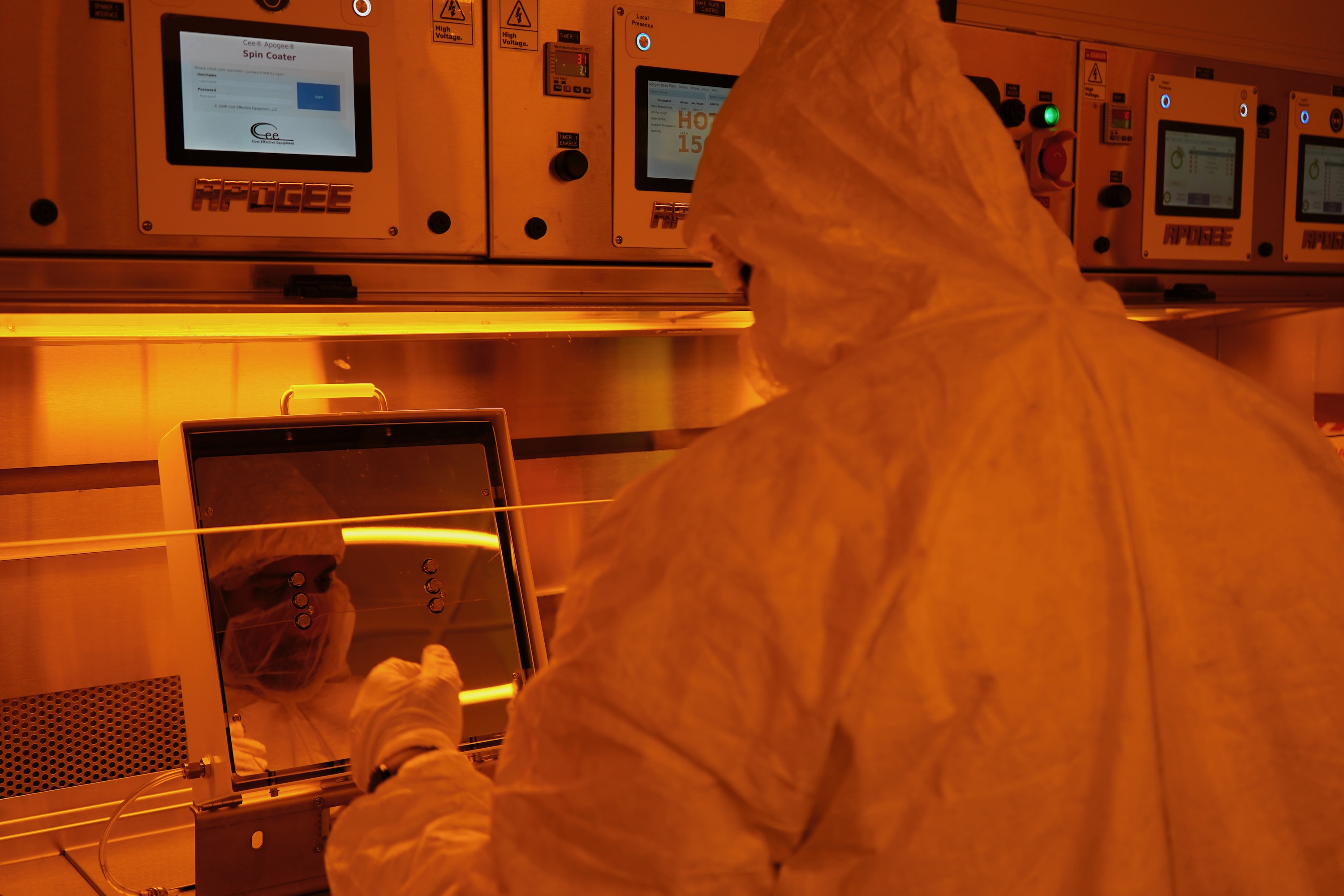 A technician in a white hazmat suit works inside an amber-lit cleanroom.