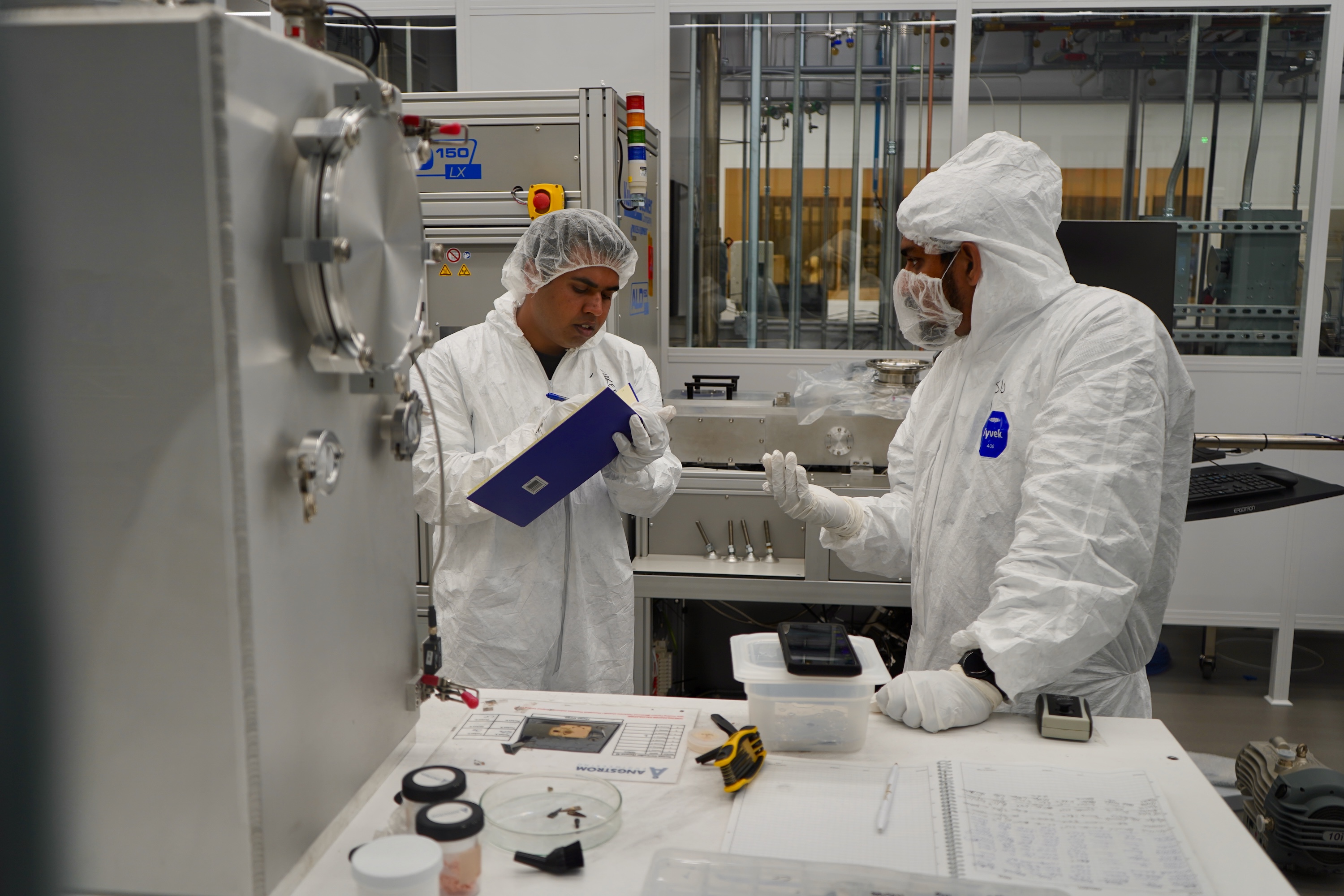 Photograph of two researchers in cleanroom suits inspecting equipment in a lab.