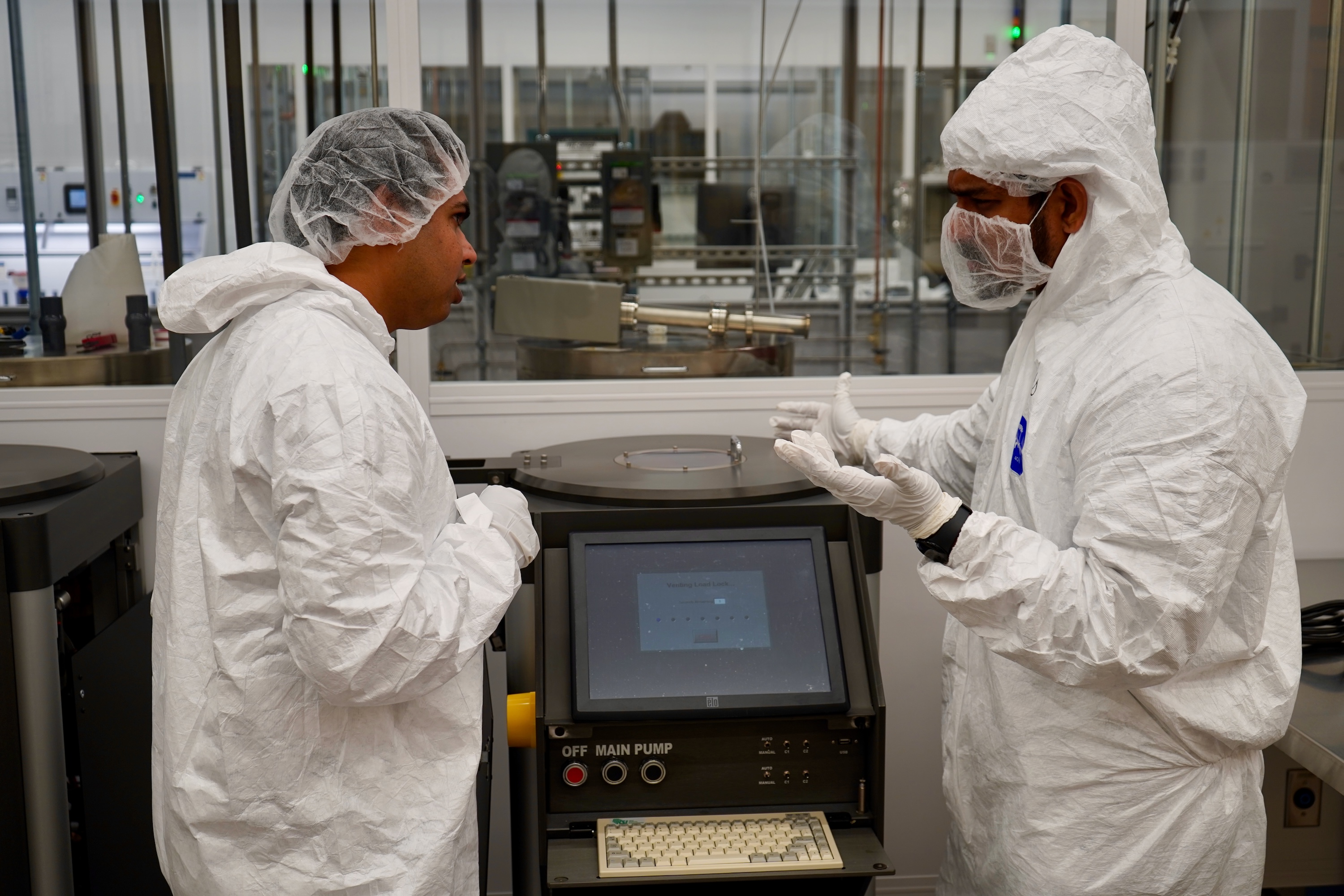 Two researchers in white cleanroom suits discuss by a machine with a monitor.