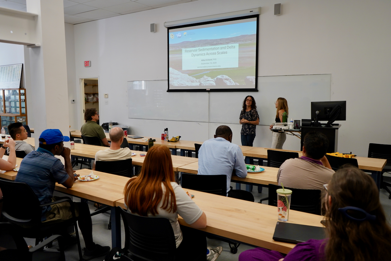 Photograph of an audience seated at tables in a room watching a presentation on a screen.