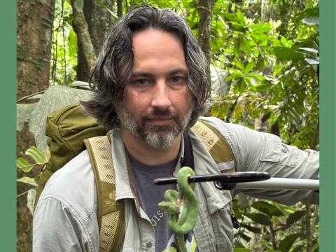 Photograph of a man in a jungle holding a small green snake on a pole.