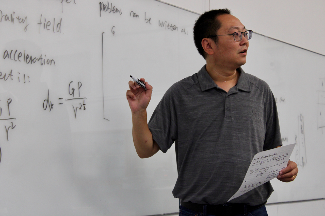 Photograph of a man in a gray polo shirt pointing at a whiteboard with math formulas.